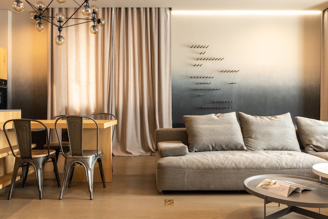 Comfortable couch with cushions and wooden table with chairs placed in modern living room with decorated wall and curtains at home